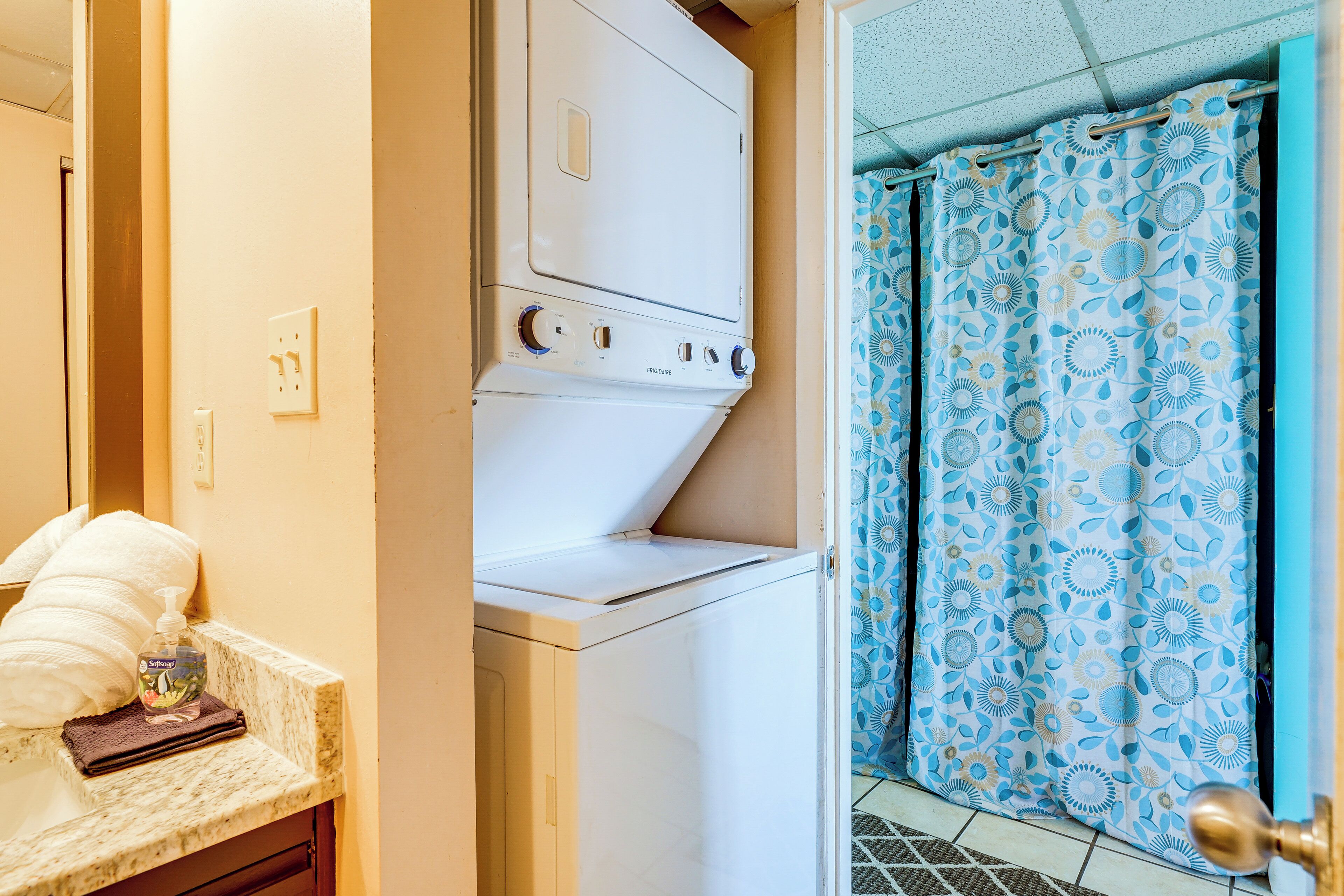 Un espace buanderie équipé d’un lave-linge et d’un sèche-linge à chargement frontal, d’un porte-serviettes et d’une salle de bain avec un rideau de douche à motifs.