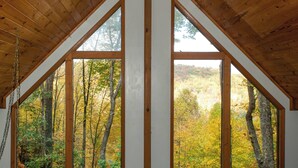 A room with a sloped wooden ceiling and large windows offering a view of a forest with autumn foliage. - Mountain-view Robbinsville Cabin w/ Hot Tub! (Robbinsville)