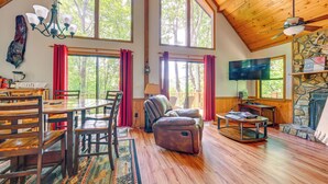 A spacious living room with wooden floors, a stone fireplace, and large windows offering a view of trees. - Mountain-view Robbinsville Cabin w/ Hot Tub! (Robbinsville)