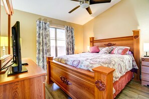 Une chambre à coucher avec un lit en bois, un ventilateur de plafond, un téléviseur posé sur un meuble en bois et une fenêtre avec des rideaux.