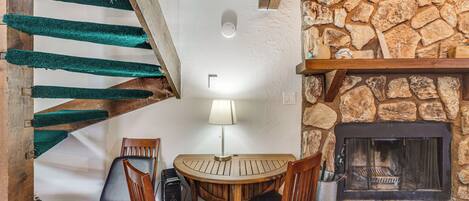 A cozy room with a stone fireplace, a wooden table with chairs, a lamp, and a staircase with green carpeted steps.