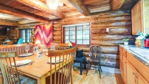 A wooden cabin interior with a dining table set for four, a kitchen area, and a large window. - Cozy Elizabethtown Cabin w/ Fire Pit! (Elizabethtown)
