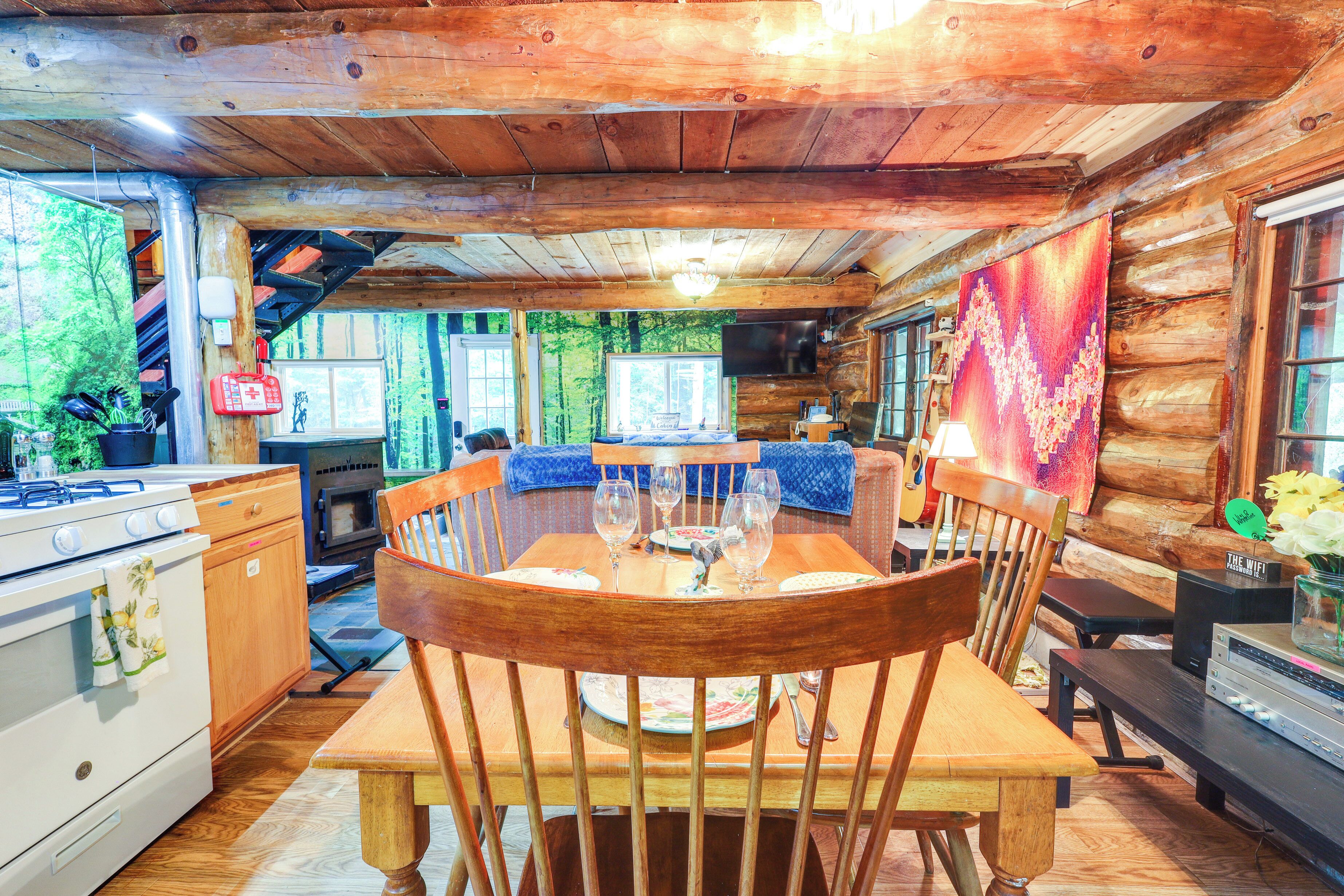 Interior rústico de una cabaña con una mesa de comedor de madera, sillas, una zona de cocina con estufa y horno, y un ventanal con vistas a un bosque.