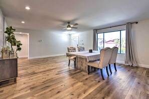 A spacious dining area with a wooden table and upholstered chairs, a ceiling fan, and large windows with curtains.