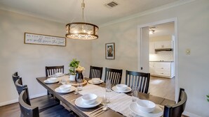 A dining room with a long wooden table set for six, surrounded by dark wooden chairs. A chandelier hangs from the ceiling, and a framed inspirational quote is on the wall.