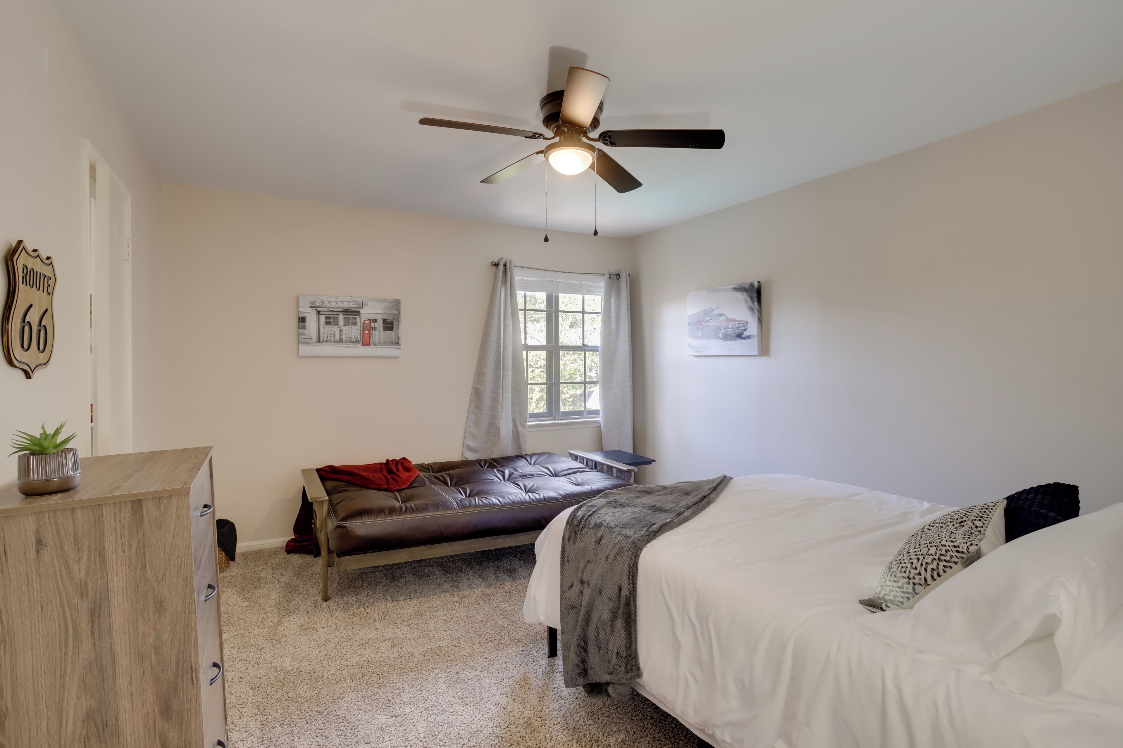 A bedroom with a bed, a bench, a ceiling fan, a window with curtains, and wall art.