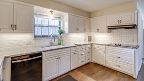 A kitchen with white cabinets, a granite countertop, a dishwasher, and a window with blinds.
