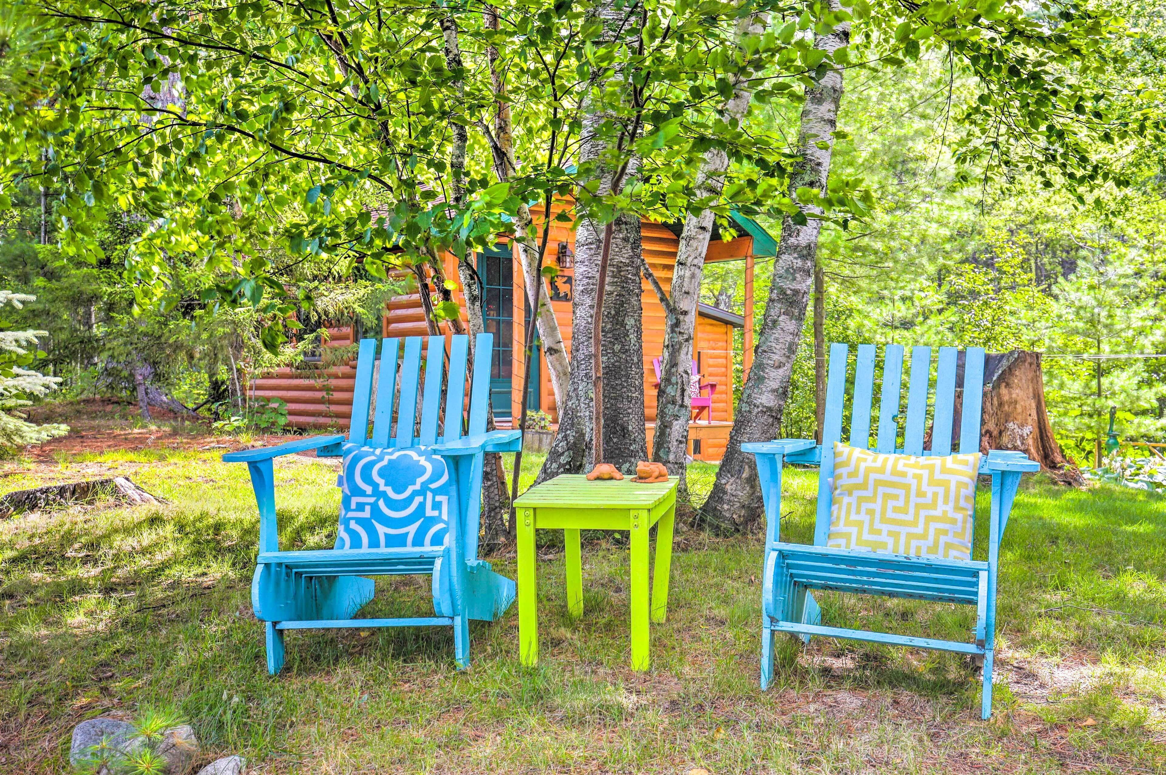 Deux chaises Adirondack bleues avec des coussins à motifs, une petite table verte et un chalet en bois situé dans une zone boisée.