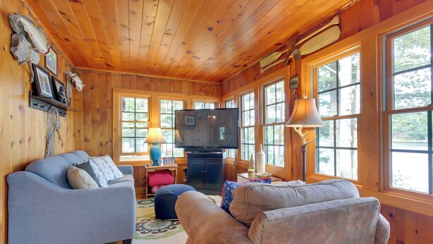 A cozy cabin interior with wooden walls and floors, a gray sofa, a blue ottoman, and a television in the center. - Lakefront Cabin w/ Private Dock & Fire Pit! (Minocqua)