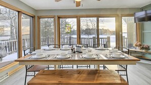 Uma sala de jantar com uma mesa grande posta para quatro pessoas, um ventilador de teto e grandes janelas com vista para um deck coberto de neve.