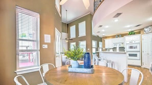 A dining area with a round wooden table, white chairs, a window with blinds, and a kitchen in the background. - Beautiful Montgomery Home w/ Lakefront Deck! (Montgomery)