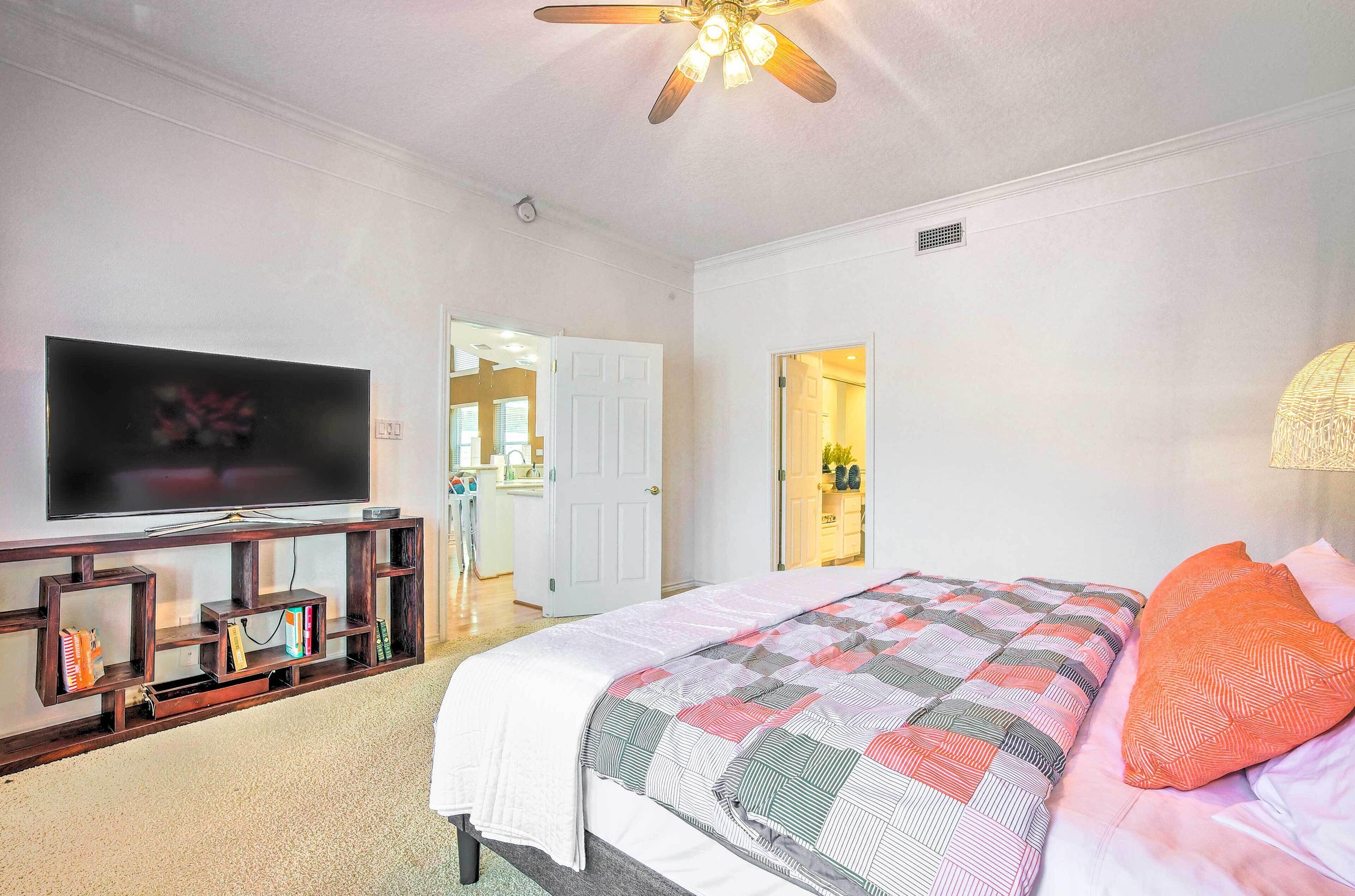 A bedroom with a large flat-screen TV, a bed with a patterned blanket, a ceiling fan, and a door leading to a bathroom.