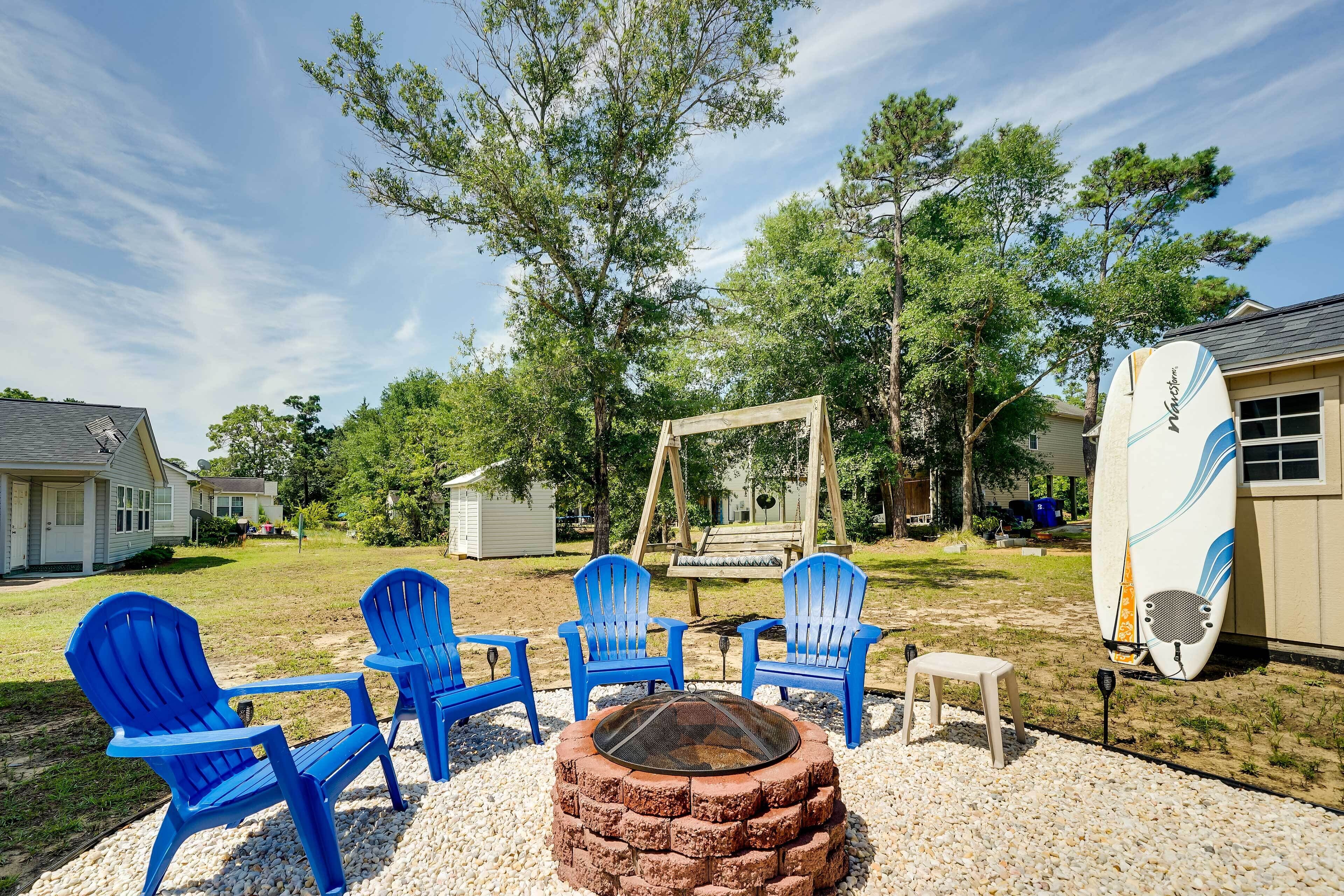 A backyard with a fire pit, blue Adirondack chairs, and a surfboard leaning against a shed.