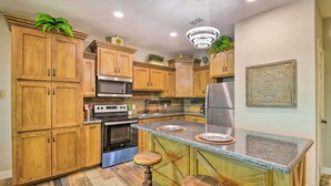Uma cozinha com armários de madeira, uma ilha central com bancos, eletrodomésticos de aço inoxidável e plantas decorativas.