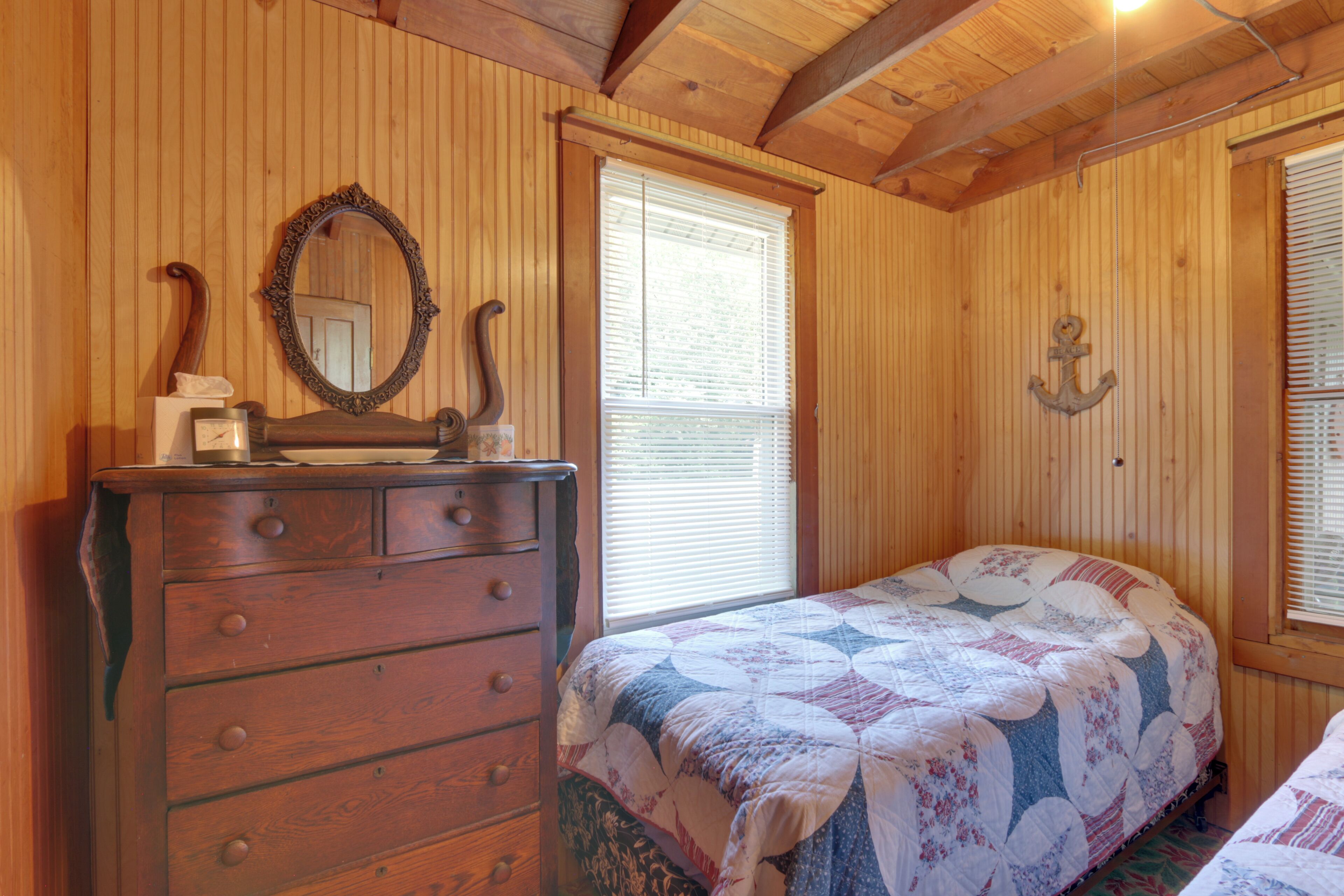 Une chambre à coucher aux murs lambrissés, avec une commode, un lit recouvert d’une courtepointe, un miroir et une fenêtre munie de stores.