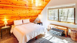 Une chambre confortable avec un plafond en pente, un grand lit, une table de chevet avec une lampe, un banc et une fenêtre donnant sur les arbres.
