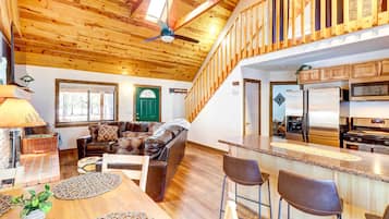 A spacious living area with a high ceiling, wooden beams, a green door, a brown leather sofa, a wooden dining table, and a kitchen with stainless steel appliances.