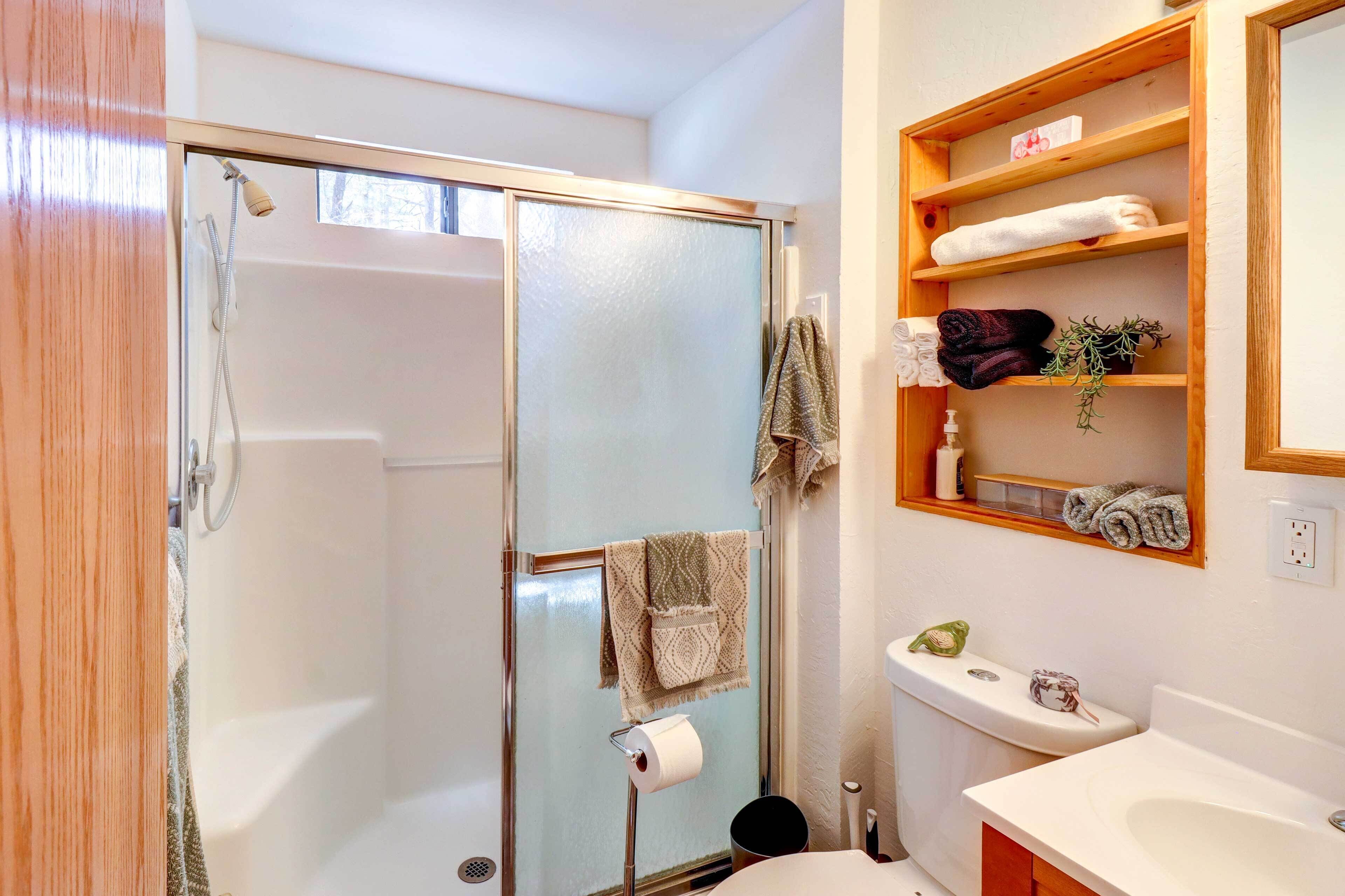 A bathroom with a glass shower enclosure, a toilet, a sink, and a shelf with towels and toiletries.