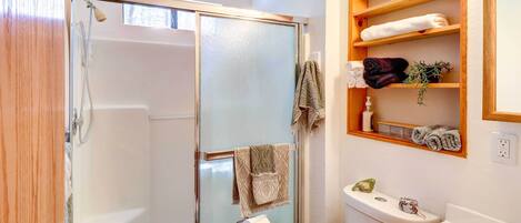 A bathroom with a glass shower enclosure, a toilet, a sink, and a shelf with towels and toiletries.