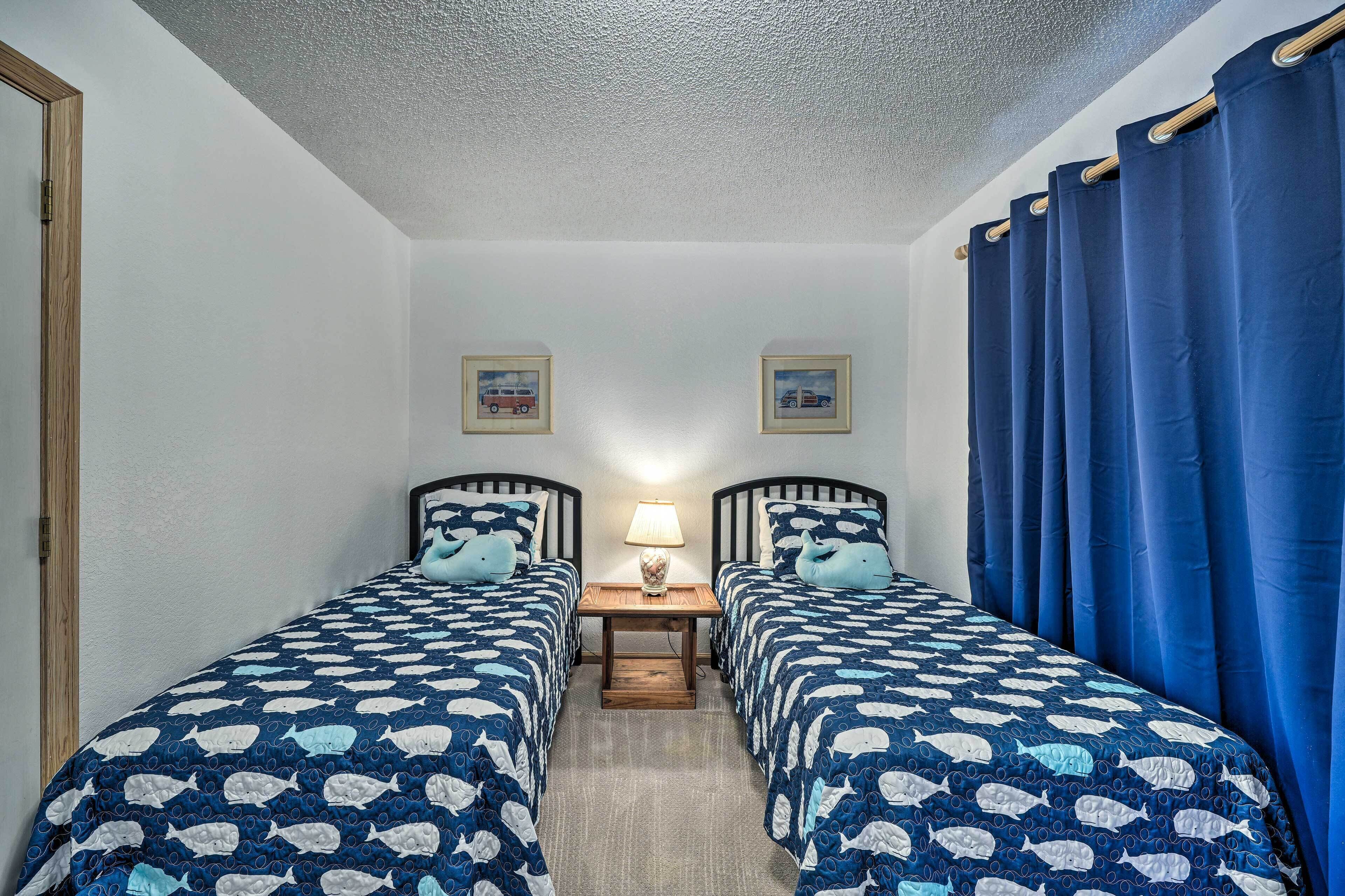 A room with two beds, each with matching blue and white bedding, a small table with a lamp, and a picture frame on the wall.