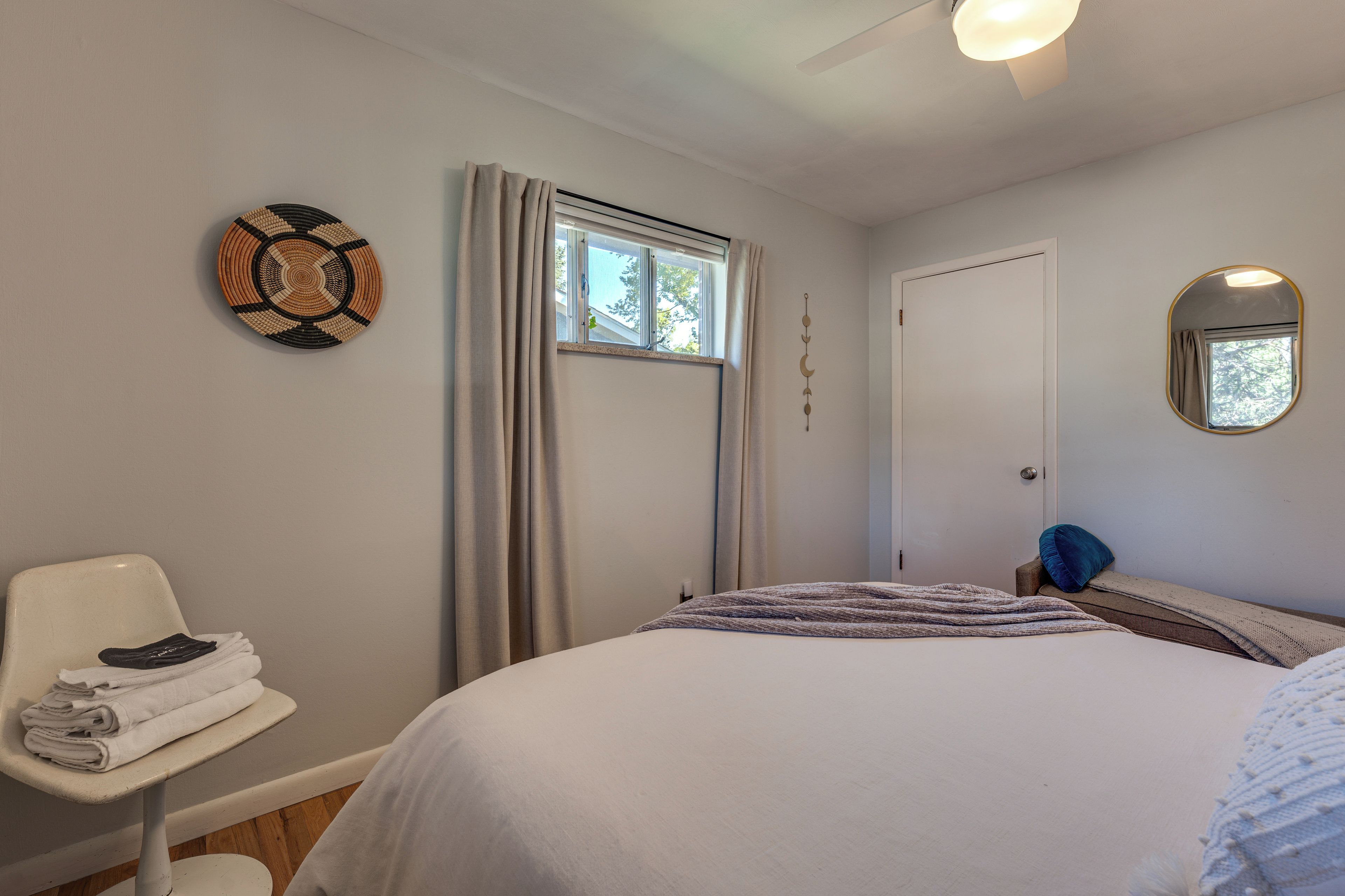 A bedroom with a bed, a chair, a window with curtains, a door, and a wall-mounted mirror.