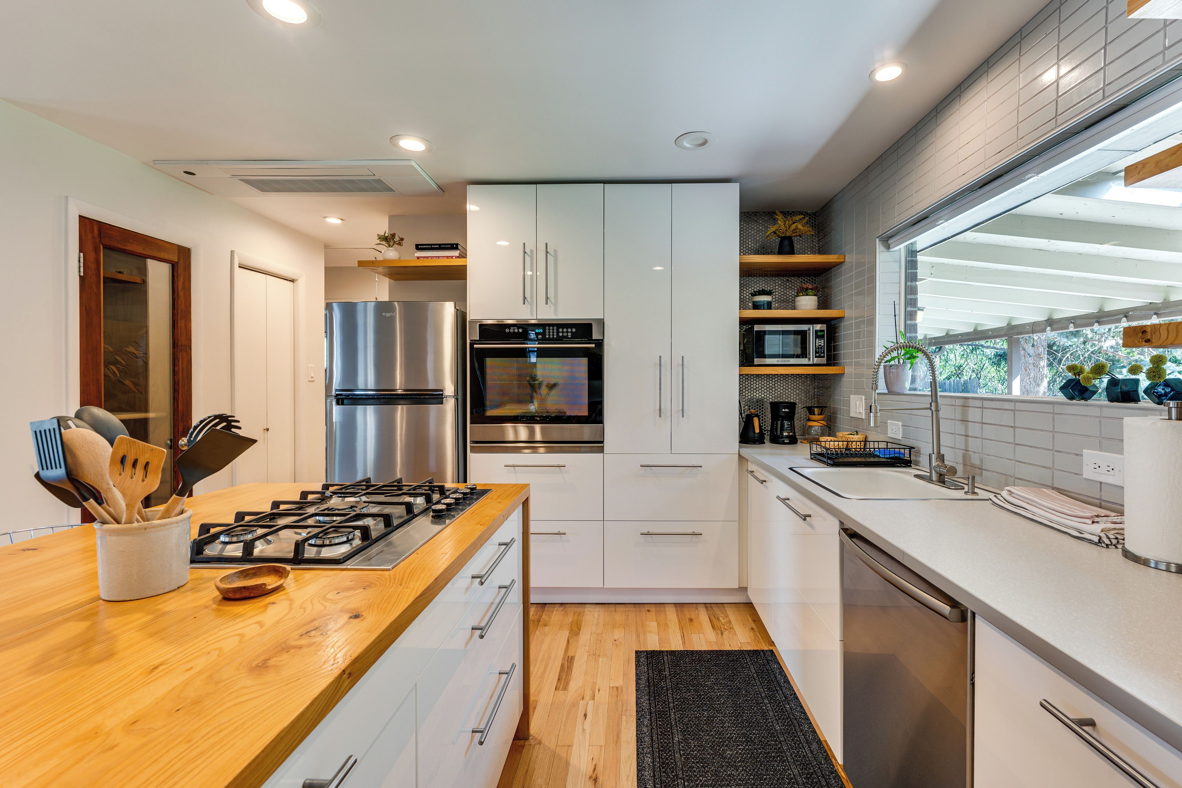A modern kitchen with a wooden countertop, stainless steel appliances, white cabinetry, and a window providing natural light.
