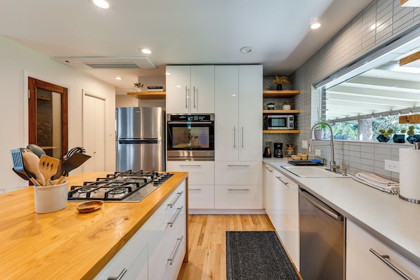 A modern kitchen with a wooden countertop, stainless steel appliances, white cabinetry, and a window providing natural light.