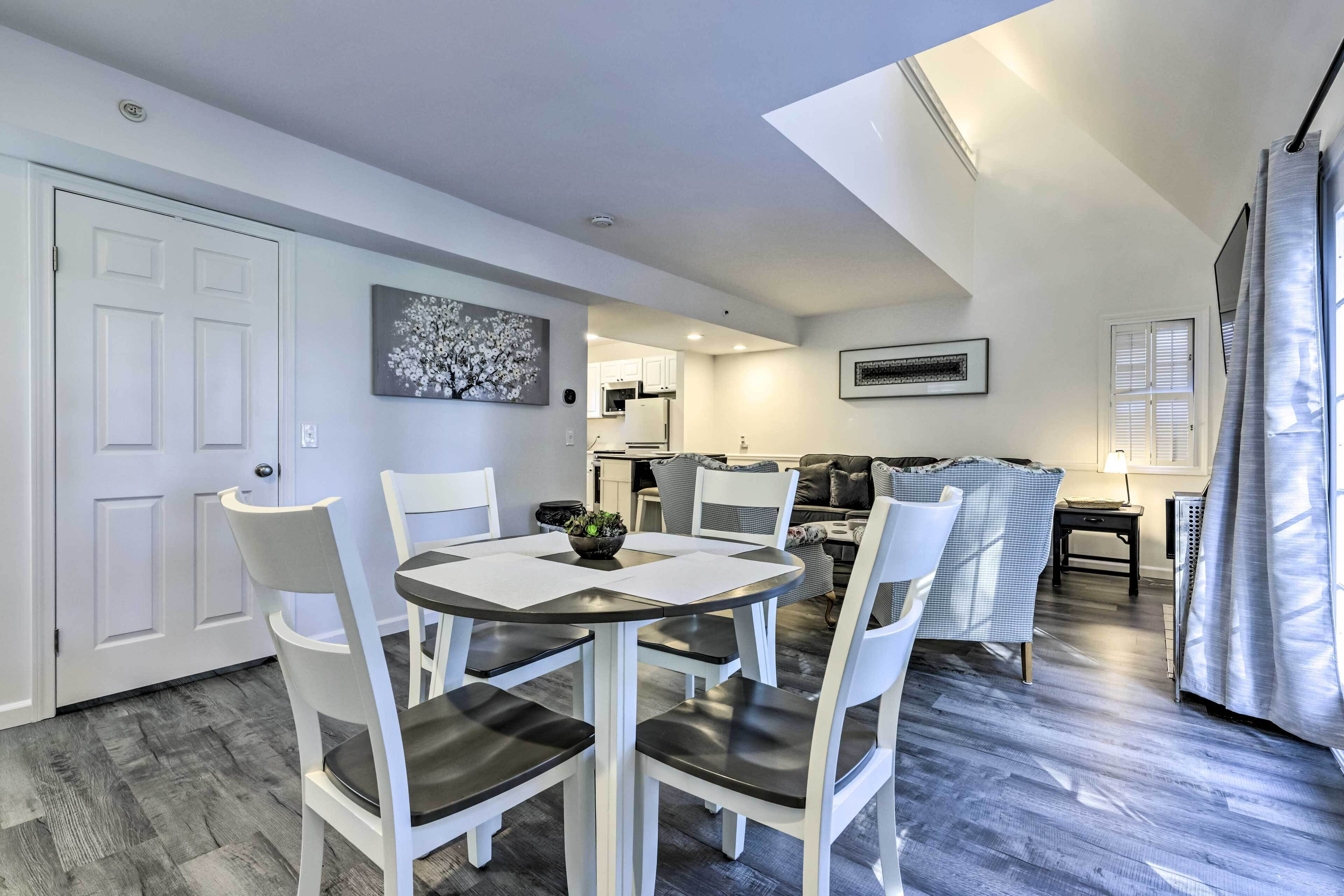 A dining area with a round table and white chairs, a kitchen in the background, and a ceiling skylight.
