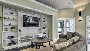 A living room with a large flat-screen TV mounted on a white entertainment unit, a gray sofa with patterned cushions, and a small round coffee table.