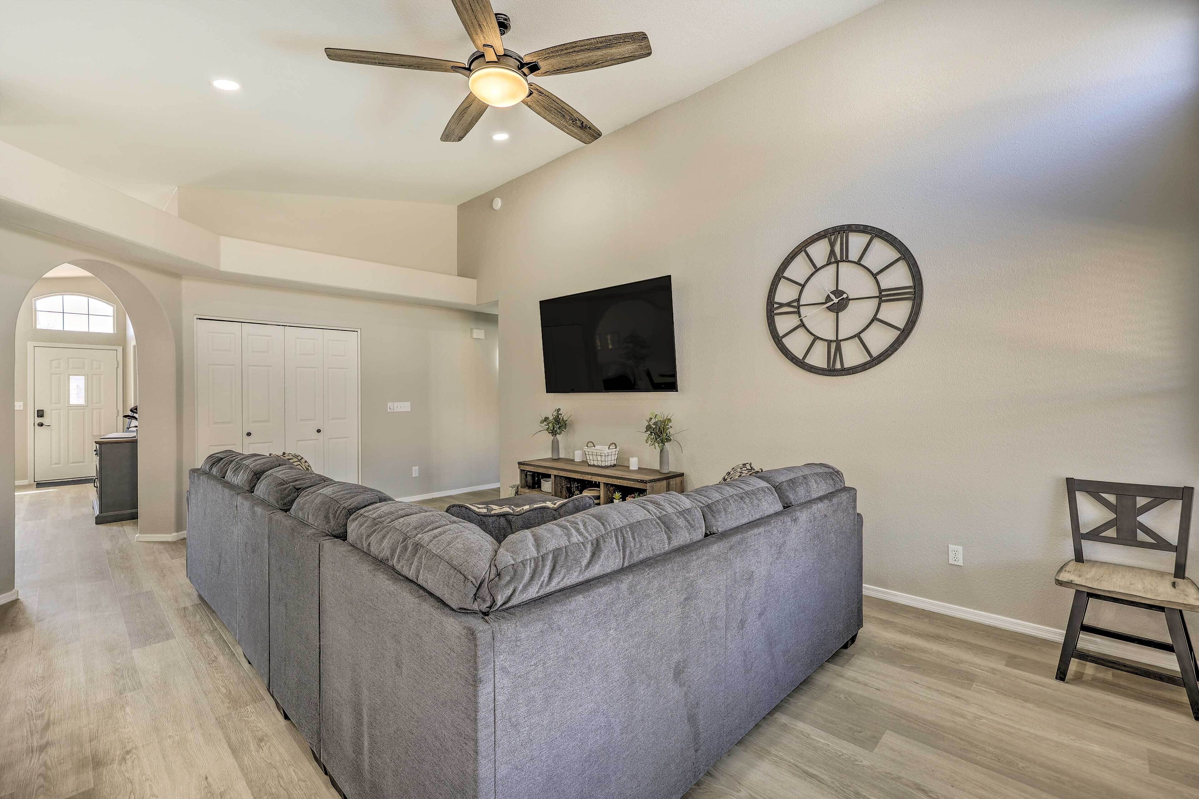 A spacious living room with a large gray sectional sofa, a ceiling fan, a wall clock, and a wooden floor.