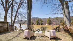 Un patio con dos sillas y una mesa, con vistas a una masa de agua y una zona boscosa.
