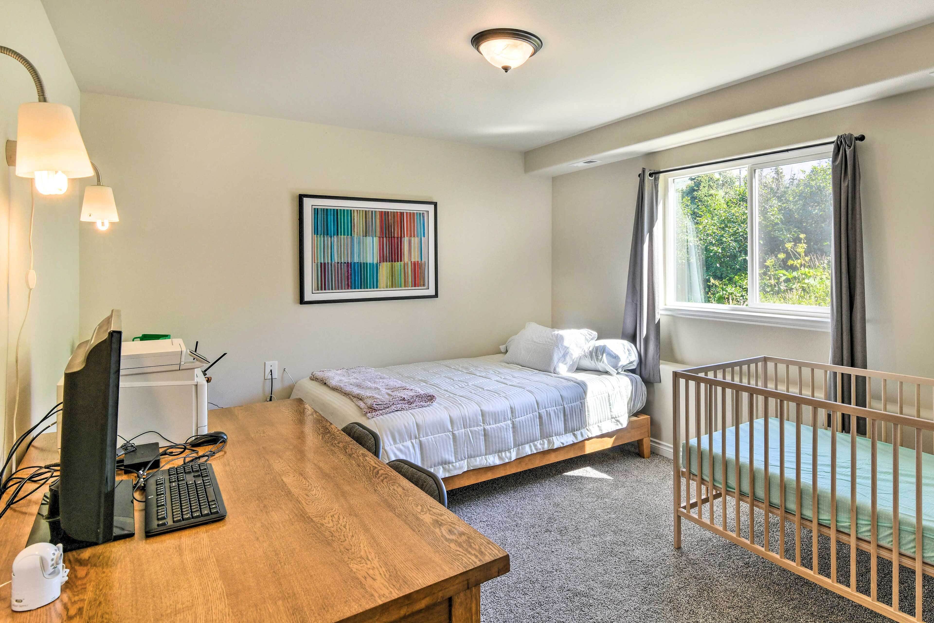 A bedroom with a bed, a desk with a computer, a crib, a window with curtains, and a colorful abstract painting on the wall.