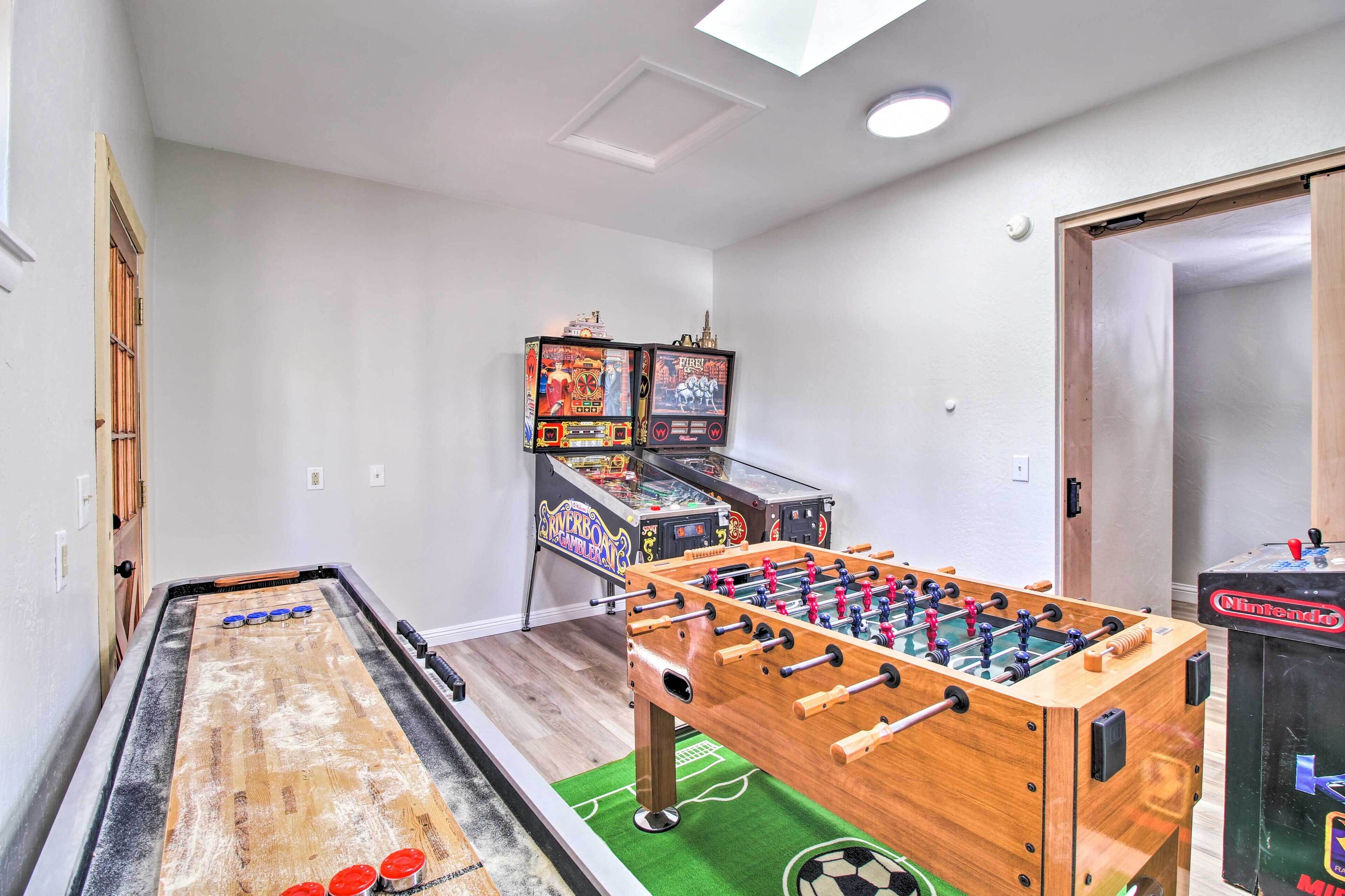 A room with a foosball table, a shuffleboard table, and two arcade machines.