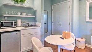 A compact kitchen with a microwave, refrigerator, and white cabinets. There is a white dining table with two chairs and a small trash bin.