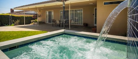 A swimming pool with a water feature, a covered patio with chairs, and a building with a tiled roof.