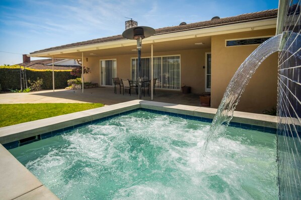 A swimming pool with a water feature, a covered patio with chairs, and a building with a tiled roof.