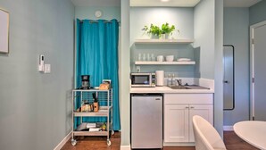 A compact kitchen with a refrigerator, microwave, and coffee maker on the counter. There is a small shelf with cups and a potted plant.