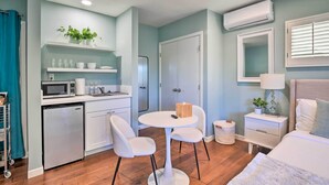 A compact hotel room with a kitchenette, a round dining table with two chairs, a bed with a white duvet, a bedside table with a lamp, and a large mirror on the wall.