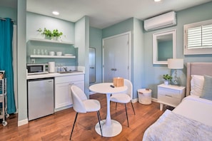 Quarto de hotel compacto com cozinha compacta, uma mesa de jantar redonda com duas cadeiras, uma cama com edredom branco, uma mesa de cabeceira com abajur e um espelho grande na parede.
