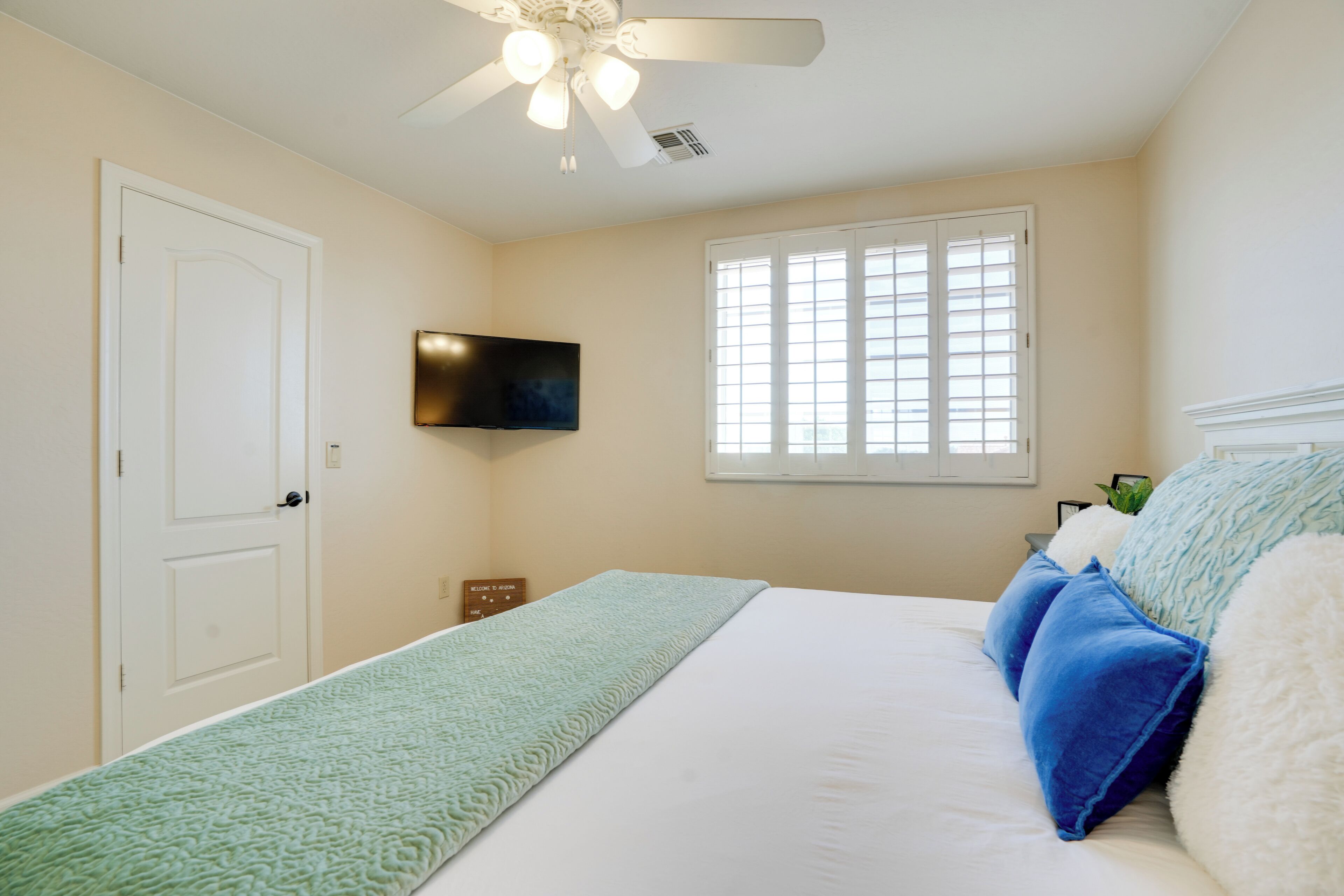 Un dormitorio con cama, ventilador de techo, televisor montado en la pared y ventana con persianas.