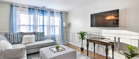 A living room with a grey sofa, patterned cushions, a coffee table with a tray, a flat-screen TV mounted on the wall, a wooden side table, and a potted plant.