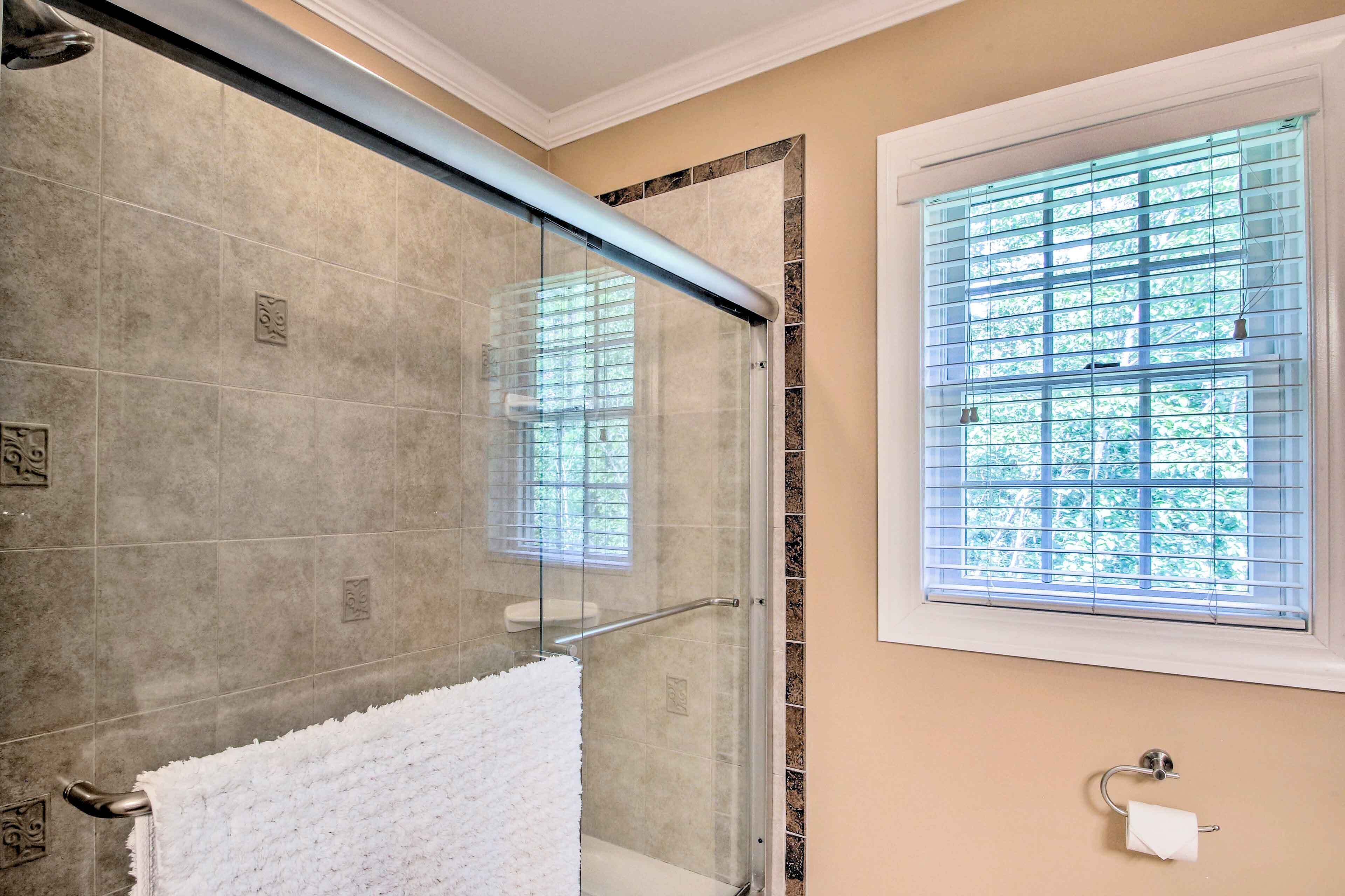 A glass-enclosed shower with a white towel, a window with blinds, and a toilet paper holder.
