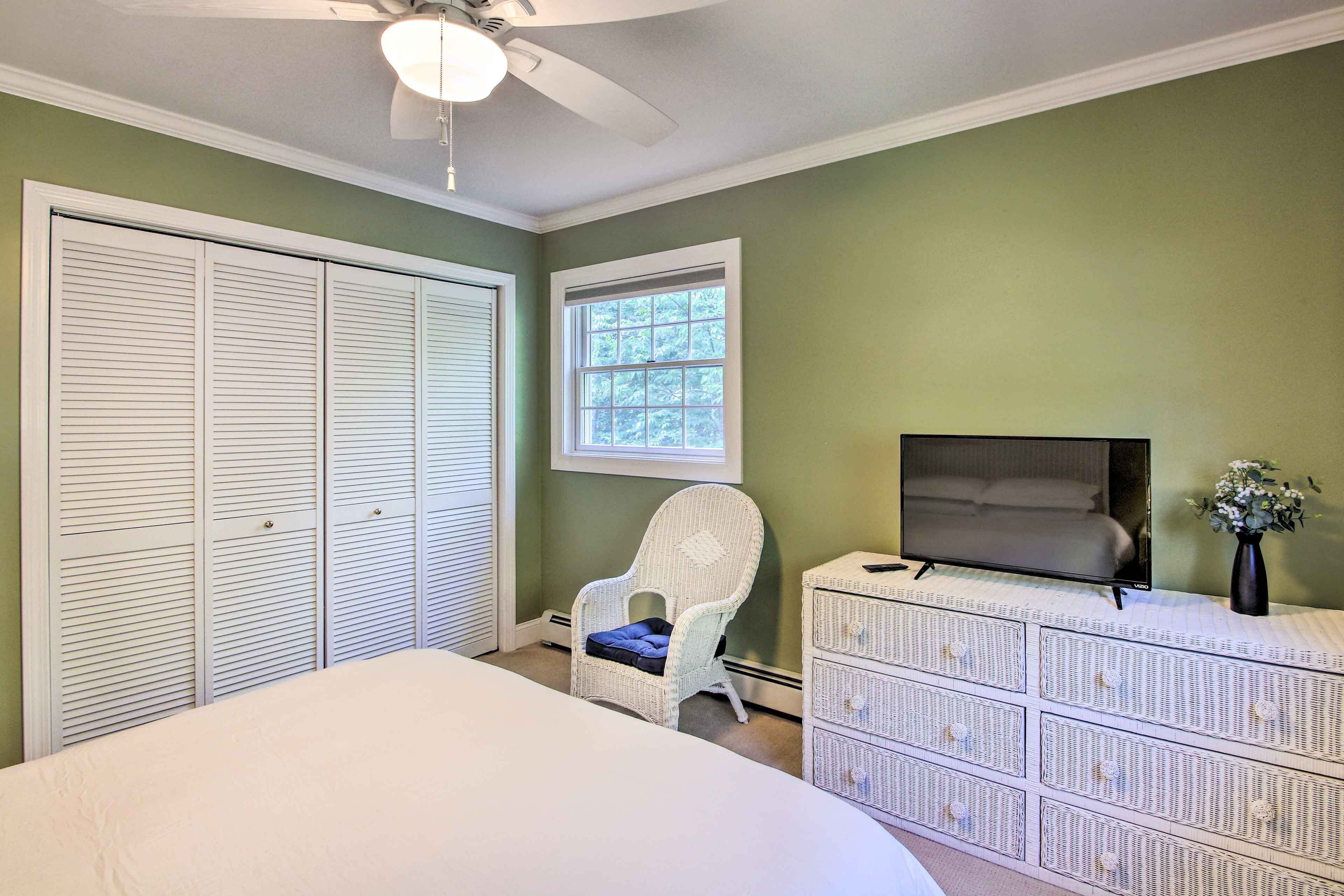 A bedroom with a bed, a white wicker chair, a dresser, a television, and a window.