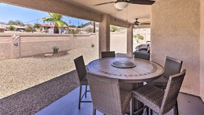 A covered patio with a round table and wicker chairs, a ceiling fan, and a view of a residential area with houses and a palm tree.