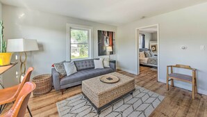 A living room with a grey sofa, a wicker ottoman, a wooden bench, and a lamp. - Breezy-chic Apartment - Steps to Geneva Lake! (Williams Bay)