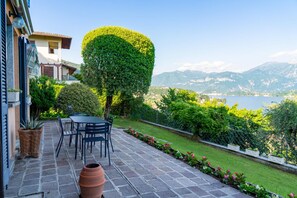 Um pátio com mesa e cadeiras, um vaso de planta e vista para um lago e montanhas.
