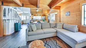 A cozy log cabin interior with a large L-shaped sectional sofa, wooden floors, and a staircase leading to an upper level.