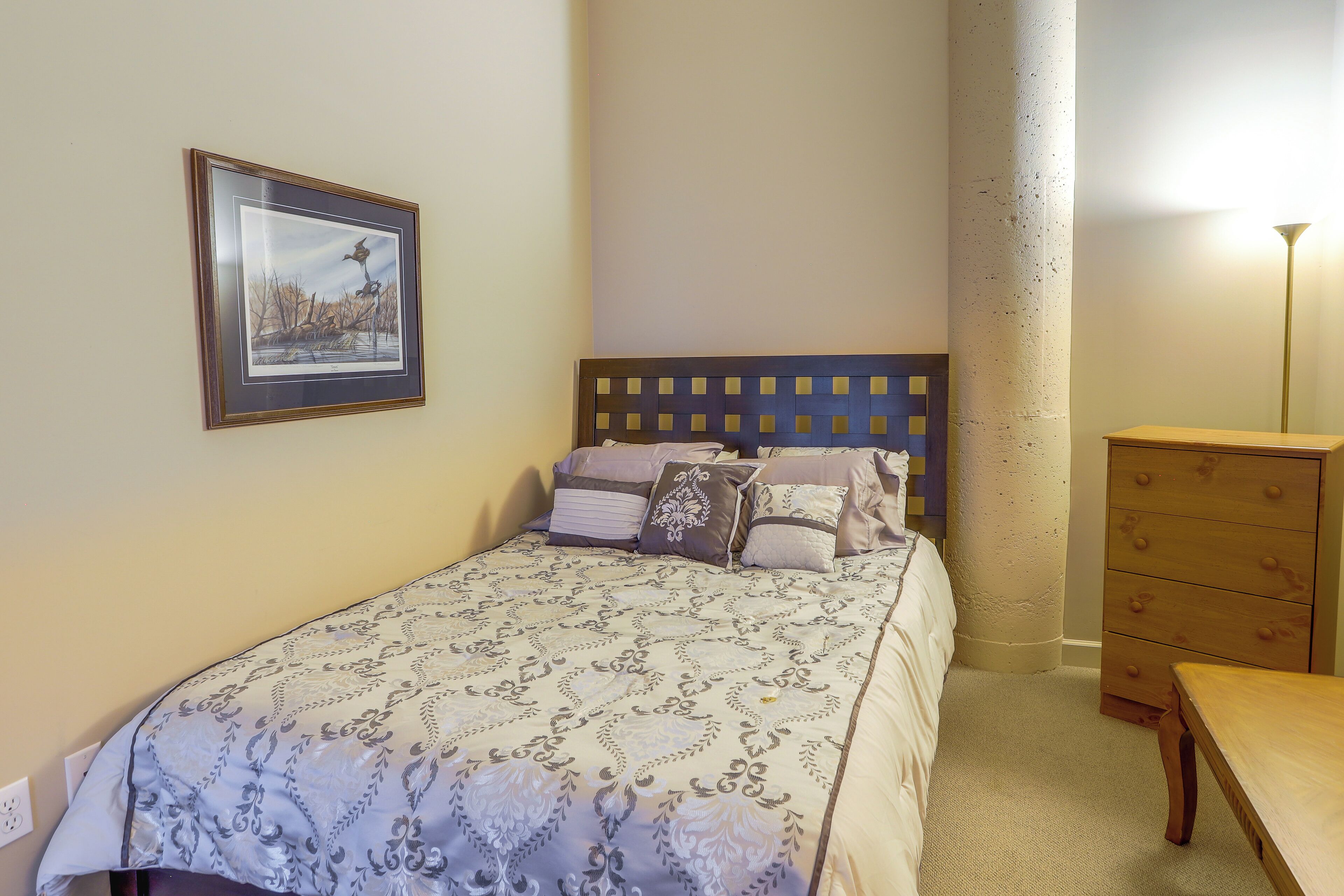 A neatly made bed with a patterned bedspread, a headboard with a grid design, a wooden dresser, and a framed picture on the wall.