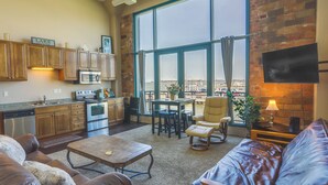 A spacious living area with a large window overlooking a body of water, a kitchen with wooden cabinets, and a leather sofa. - Waterfront Sandusky Vacation Rental: Downtown! (Sandusky)