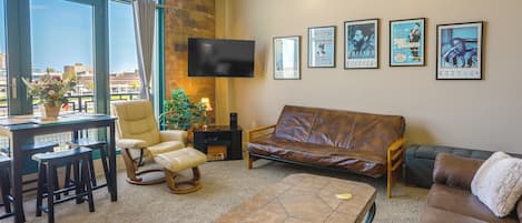 A spacious living room with a large window, a brown leather sofa, a coffee table, and a TV mounted on the wall.